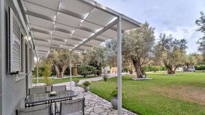 A covered patio with a stone floor, outdoor seating, and a view of a well-maintained garden with trees and shrubs. - Studio Apartment 'Elaia  3' with Mountain View, Wi-Fi and Air Conditioning (?e???da)
