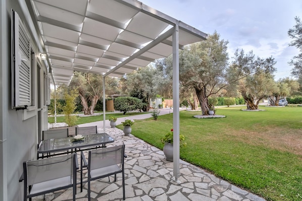 A covered patio with a stone floor, outdoor seating, and a view of a well-maintained garden with trees and shrubs.