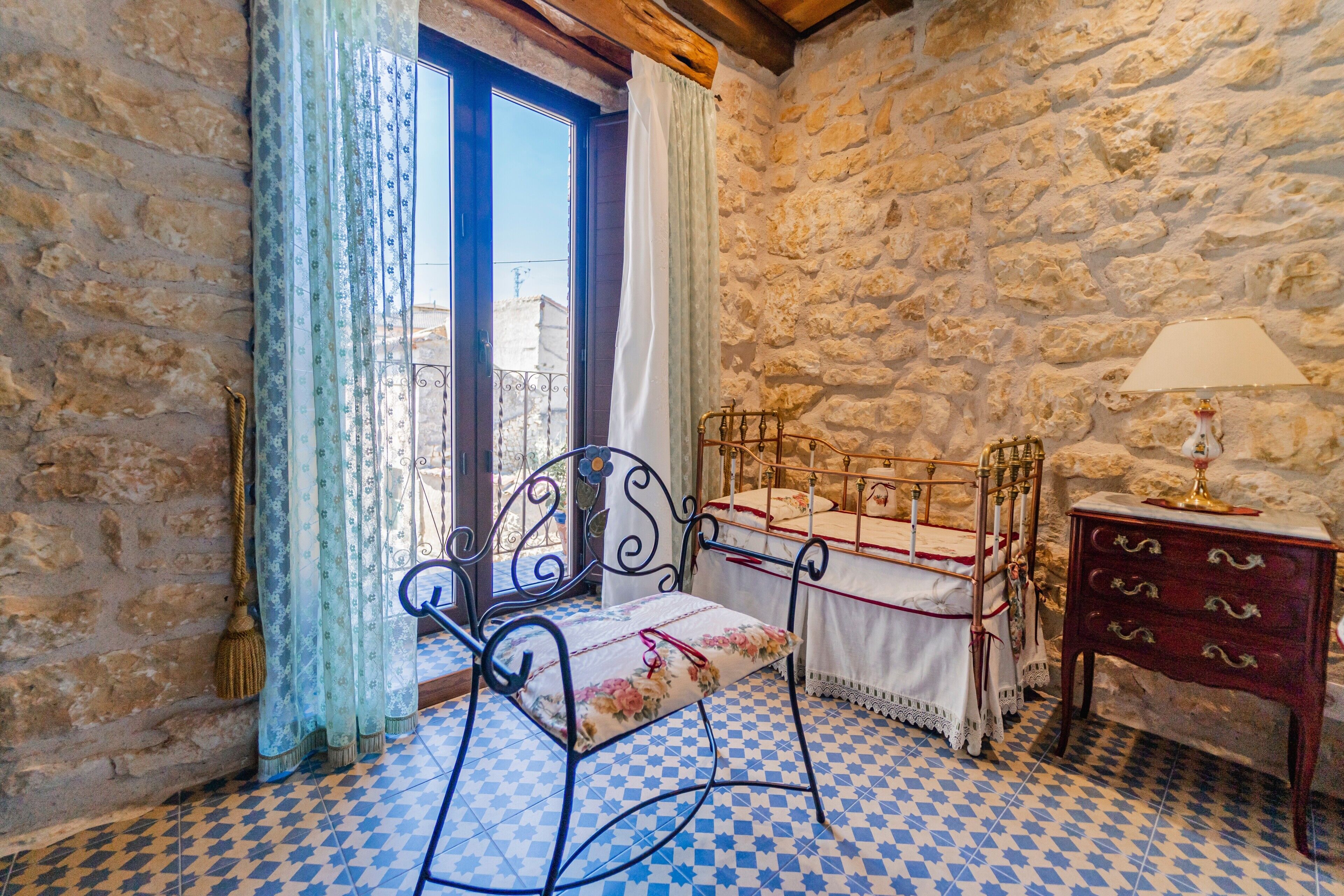 Una habitación con pared de piedra, piso a cuadros, estructura de cama de metal, mesita de noche de madera y ventana con cortinas.