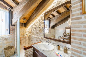 Ein rustikales Badezimmer mit Steinwand, Holzbalken, einem großen Spiegel und einem weißen Waschbecken.