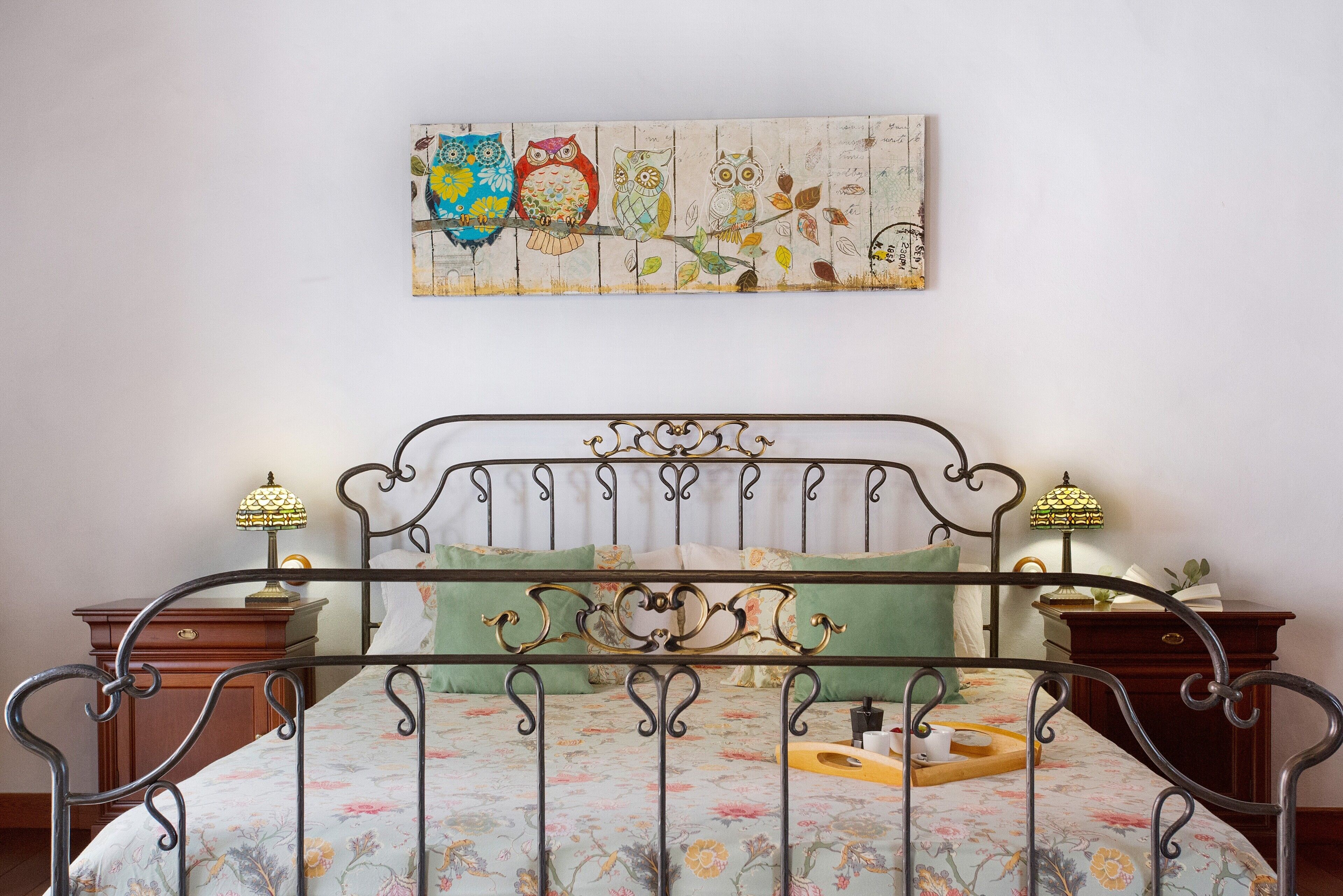 Ein Bett mit Metallgestell, blumigem Bettzeug, zwei Nachttische mit Lampen und ein an der Wand hängendes Eulenbild.