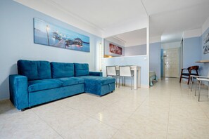 A living room with a blue sectional sofa, a dining table with chairs, and a kitchen area.