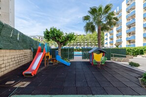 A playground with a red slide and blue climbing structures, surrounded by a paved area and a tall building in the background.
