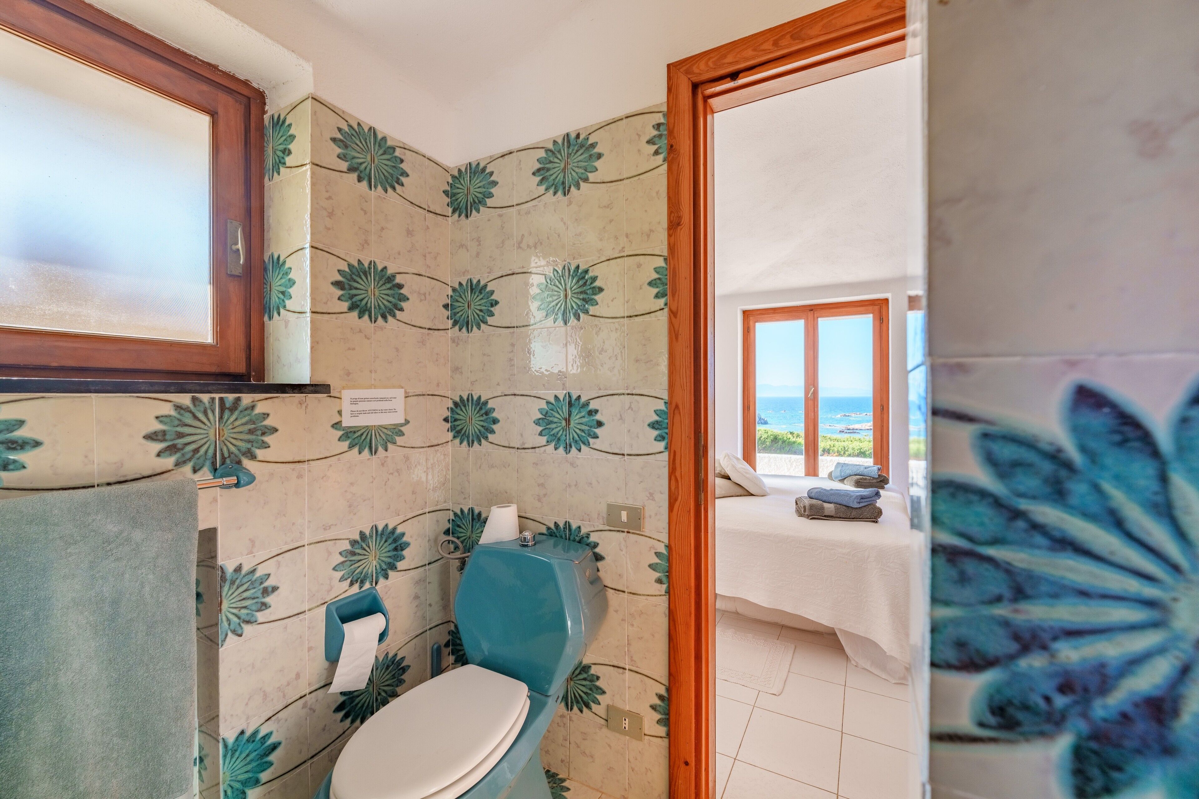 A bathroom with a toilet, a towel, and a view of a bedroom with a bed and a window.