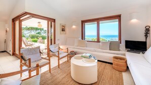 Ein geräumiger Wohnbereich mit Sofa, Sesseln, einem Couchtisch und einem Fernseher. Ein großes Fenster bietet Meerblick.