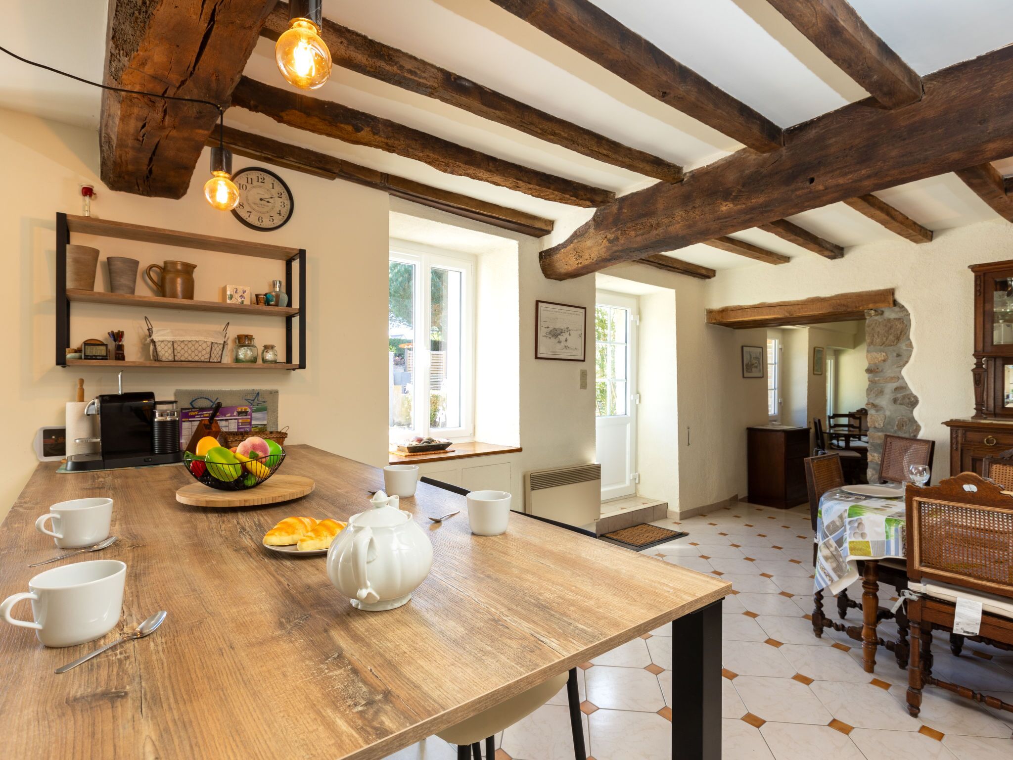 A rustic kitchen with exposed wooden beams, a wooden table with cups and a fruit bowl, and a stone fireplace.