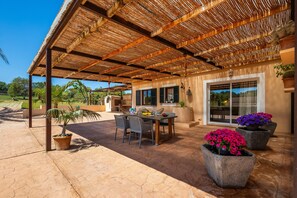 Une terrasse couverte avec une table et des chaises, des plantes en pot et un bâtiment doté de grandes fenêtres.