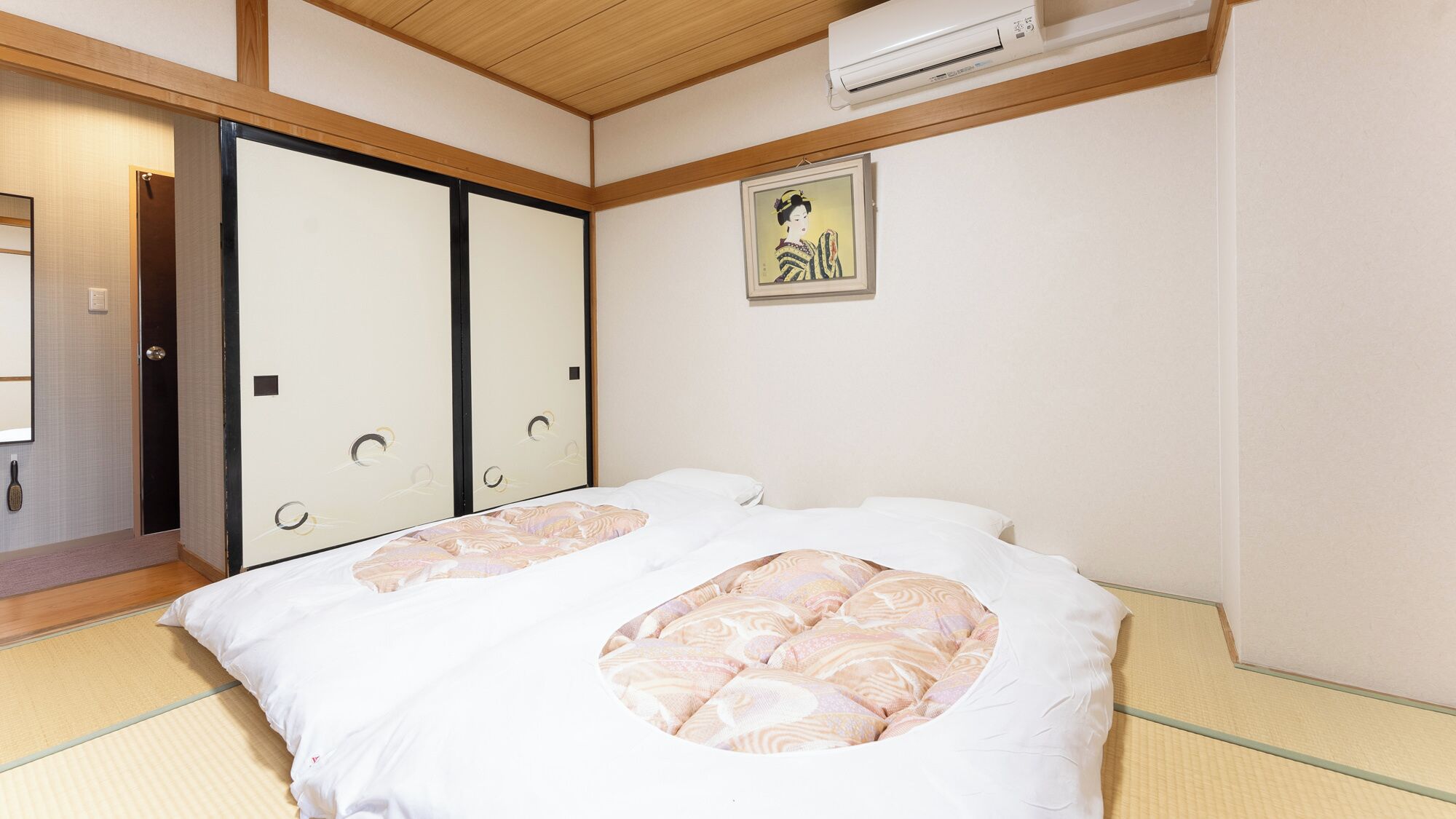 Une pièce de style japonais traditionnel, avec un sol recouvert de tatamis, un futon servant de lit, des portes coulissantes et un climatiseur fixé au mur.