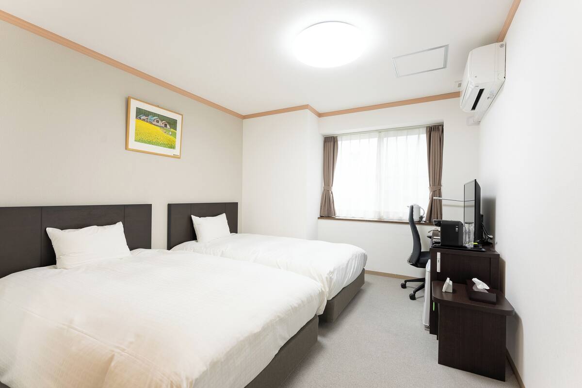 A hotel room with two beds, a desk with a computer, a chair, and a window with curtains.