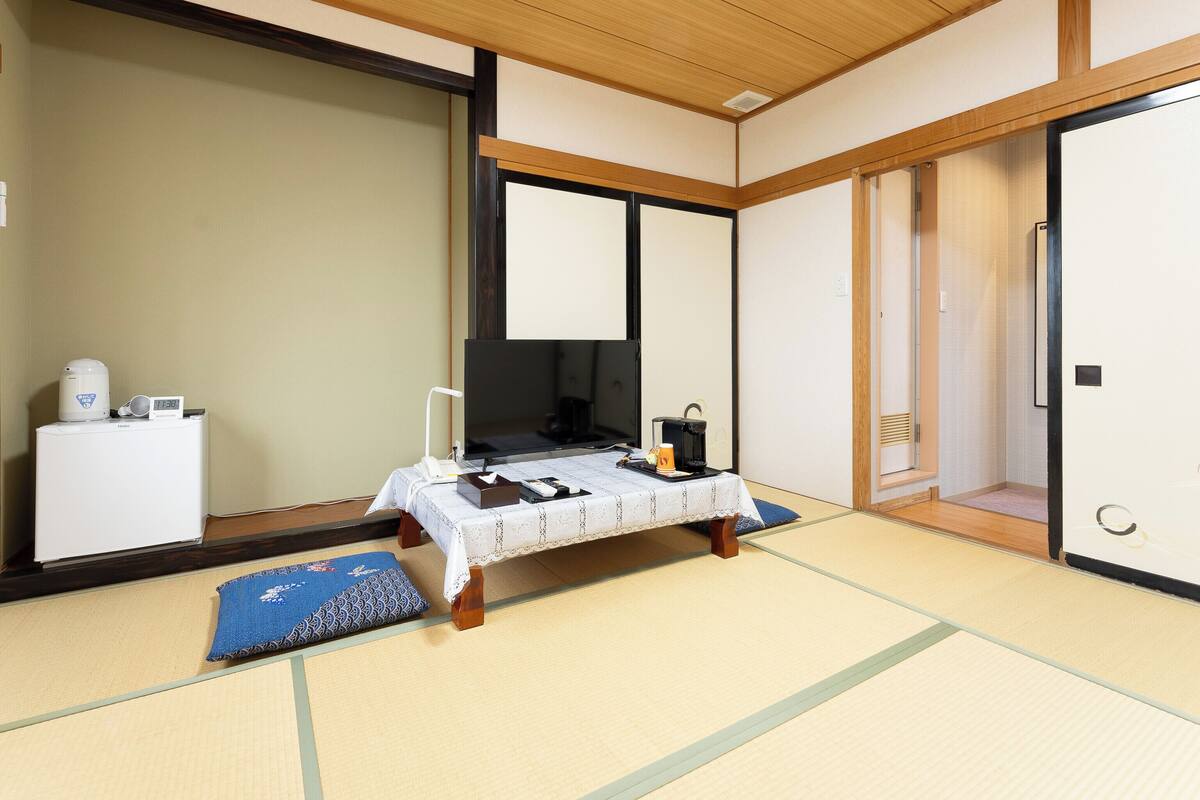 A traditional Japanese room with tatami flooring, a low table with a tablecloth, a television, a kettle, and a coffee maker.