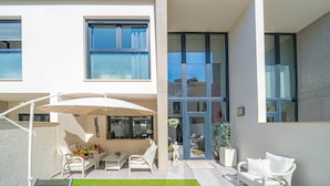 Une terrasse extérieure moderne, avec des meubles blancs, une porte-fenêtre et une vue dégagée sur l’intérieur.