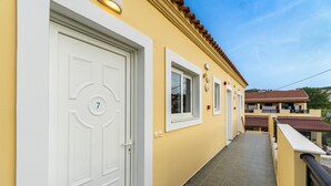 Un bâtiment jaune avec une porte blanche portant le numéro 7, un balcon carrelé et un toit recouvert de tuiles rouges.