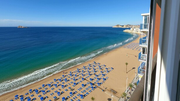 Ein Strand mit Reihen blauer Sonnenschirme und Liegen, ein klares blaues Meer und ein Gebäude mit Balkonen mit Blick auf den Strand.
