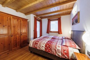 Ein Schlafzimmer mit Holzbalkendecke, einem Bett mit gemusterten Betttextilien, einem Kleiderschrank und einem Fenster mit Vorhängen.