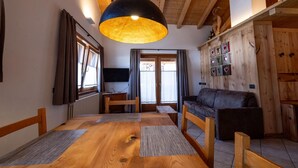A cozy living room with a wooden ceiling, a large hanging light fixture, a sofa, a TV, and a wooden cabinet with decorative items. - Apartment 'Gerry House' with Mountain View, Balcony and Wi-Fi (Livigno)