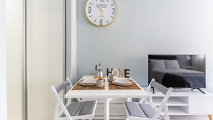 A modern room with a white table set for two, a white chair, a wall clock, a bed with a TV, and a 'HOME' sign. - Studio Apartment 'Torremolinos Dream Sea Views' with Sea View, Wi-Fi & Air Conditioning. (Torremolinos)