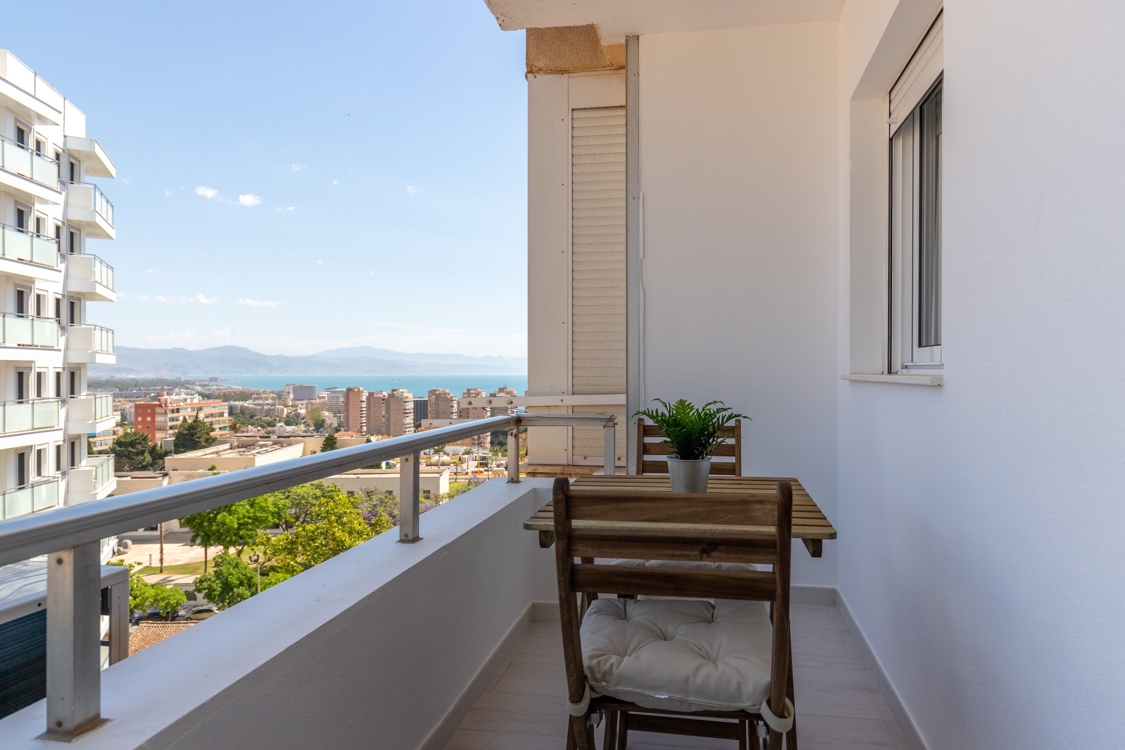 A balcony with a wooden table and chair, a potted plant, and a view of the cityscape and mountains.