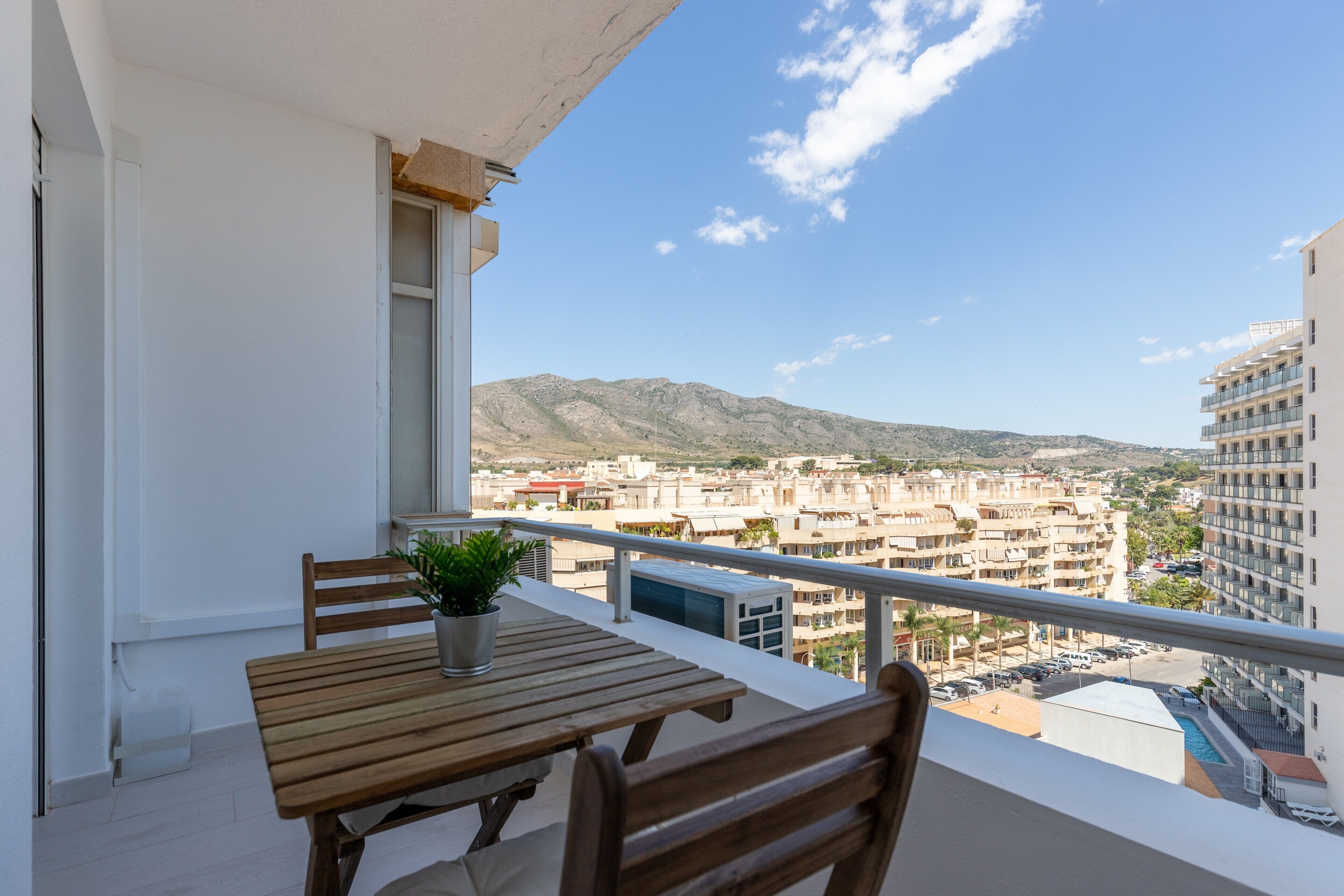 A balcony with a wooden table and chairs, a potted plant, and a view of a cityscape with buildings and mountains.