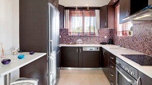 Una cocina moderna con armarios oscuros, horno integrado, lavavajillas y ventana con cortinas traslúcidas.