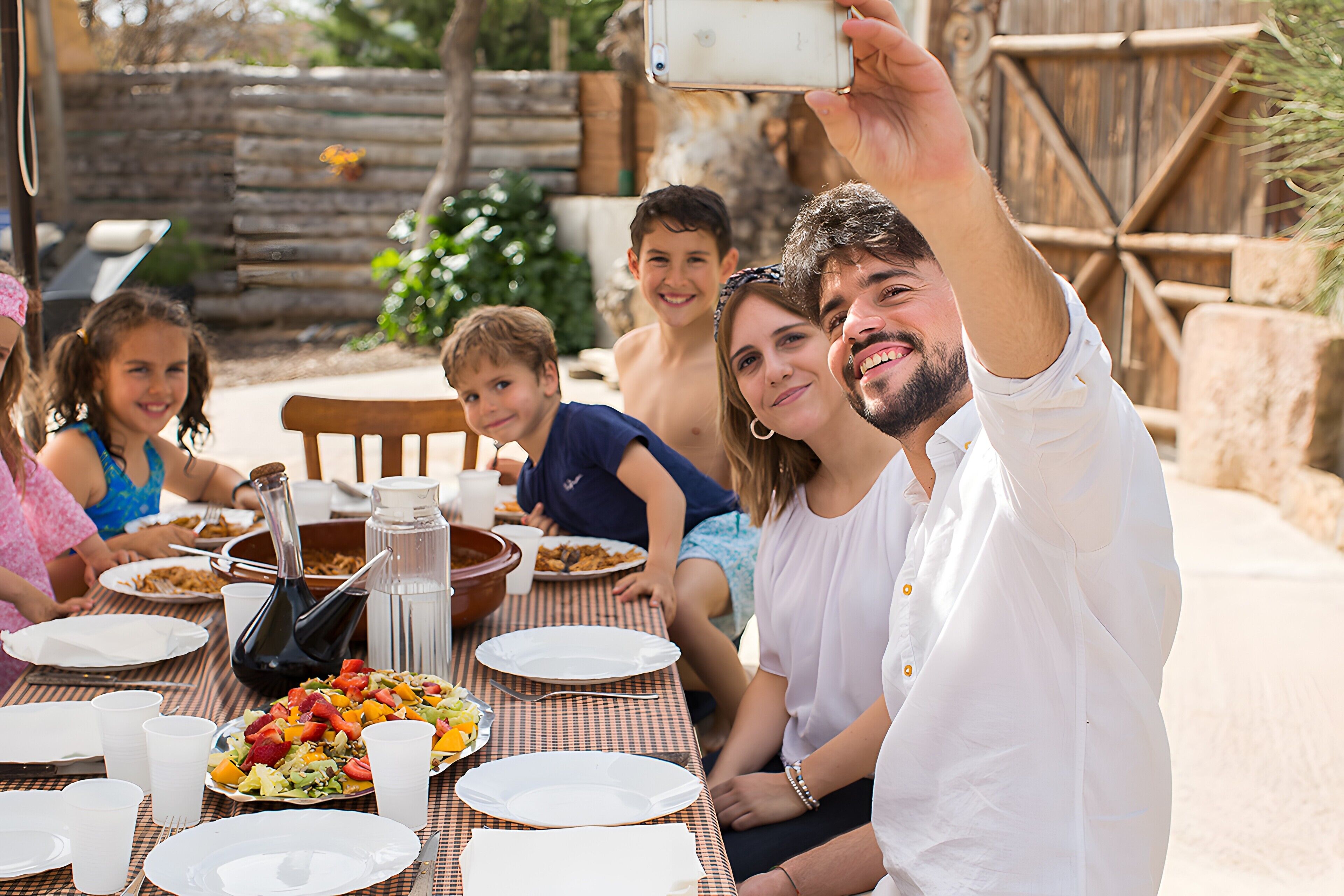 A group of people seated at an outdoor table with food, a wine bottle, and a pitcher.