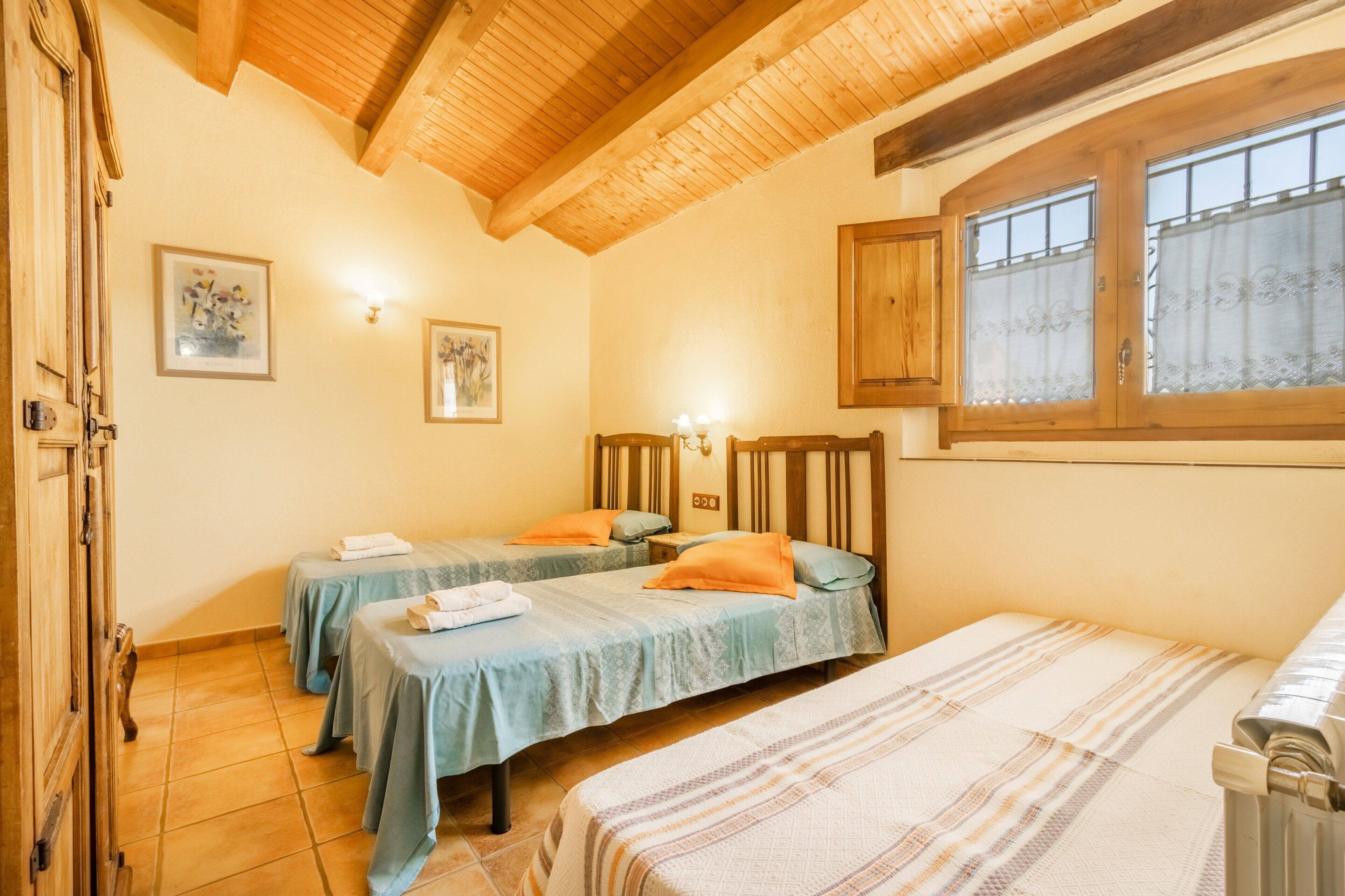 A hotel room with two single beds, a wooden ceiling, a window with curtains, and a radiator.