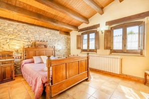 A bedroom with a stone wall, wooden ceiling beams, a wooden bed, a wooden dresser, and a window with shutters.