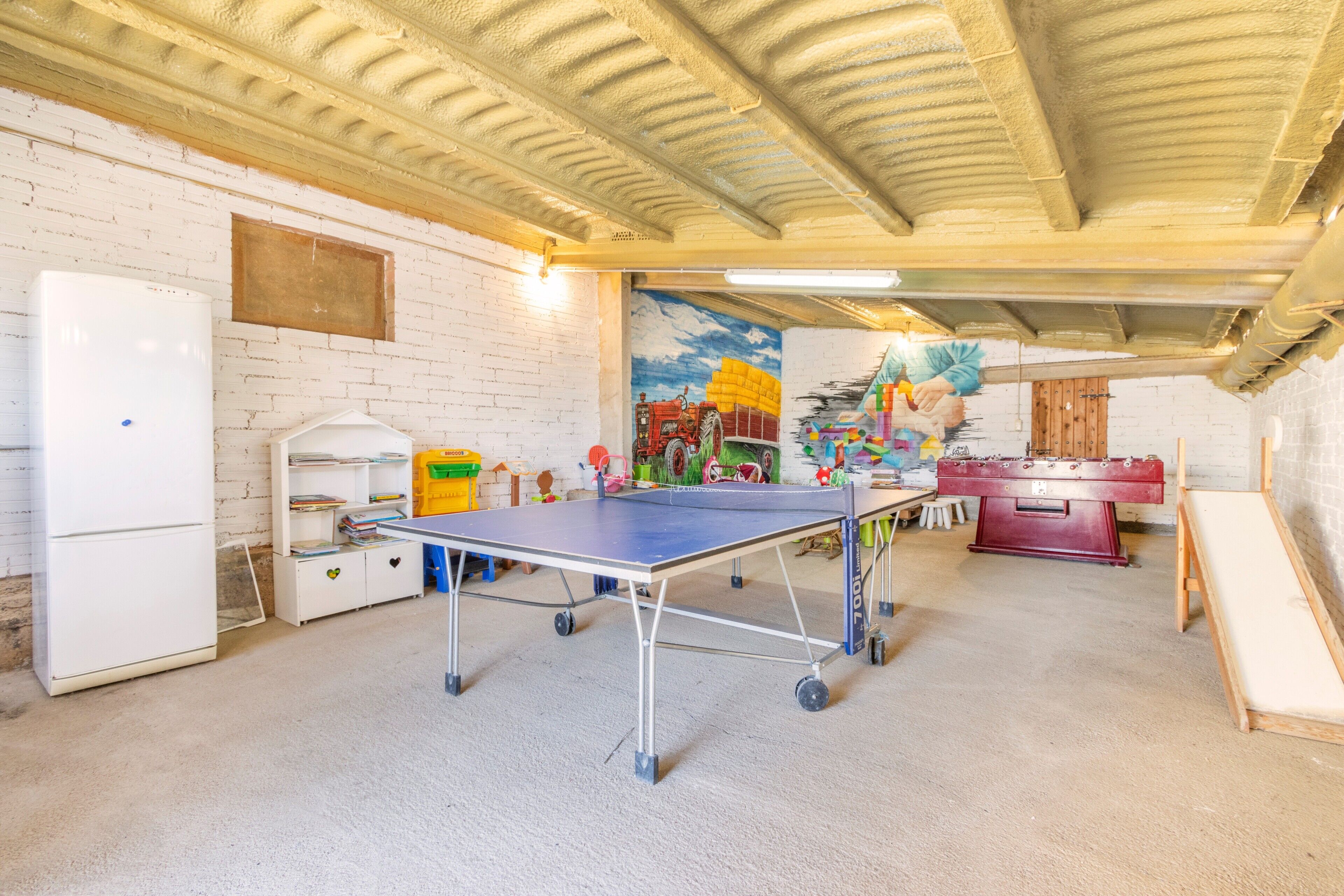 A room with a ping pong table, a refrigerator, a foosball table, and a mural on the wall.