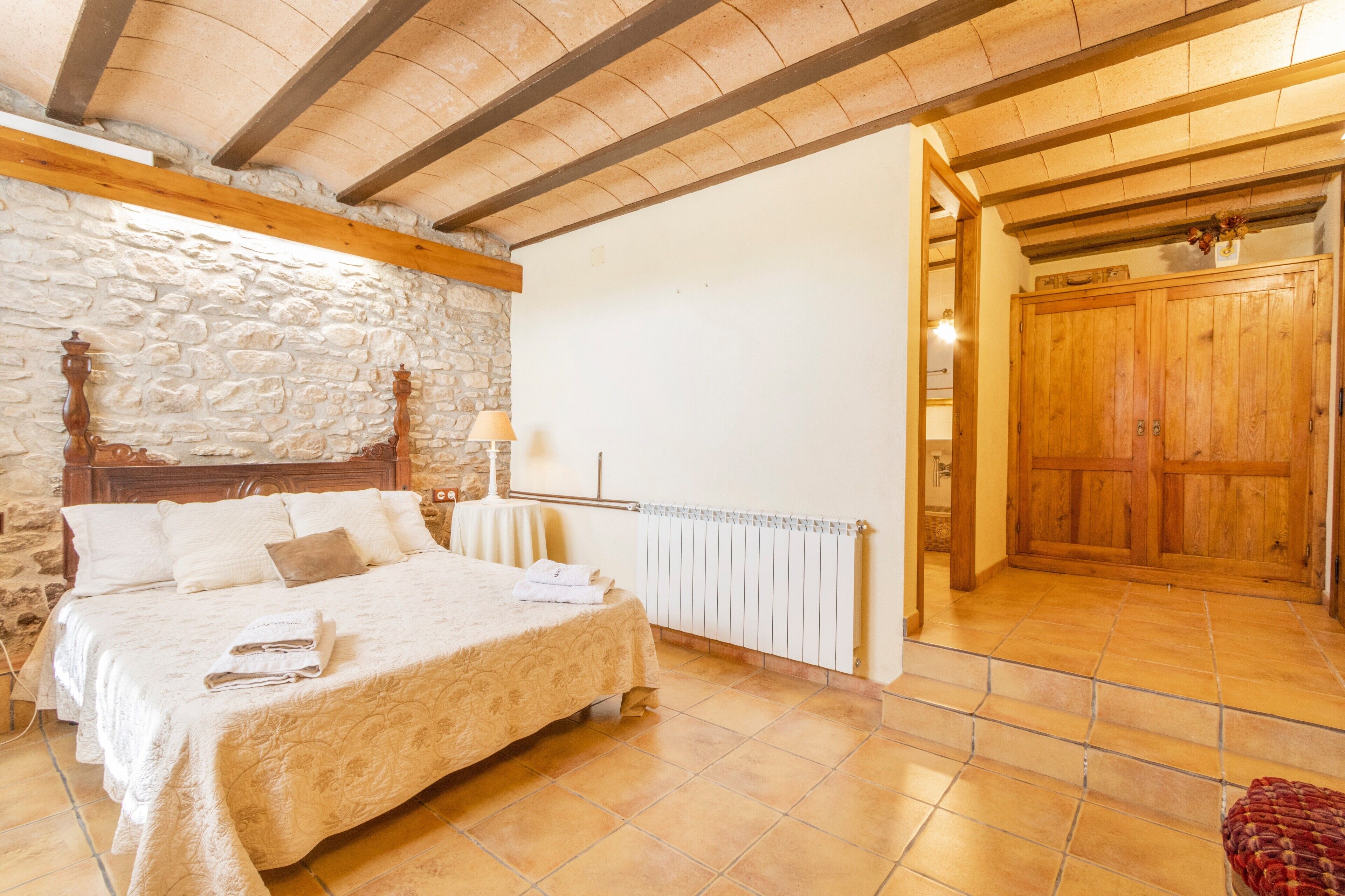 A bedroom with a stone wall, wooden ceiling beams, a bed with a quilt, a radiator, and a wooden door.