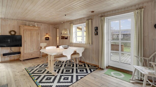 Et træinteriør med spisebord, stole, et fjernsyn og et stort vindue med gardiner.