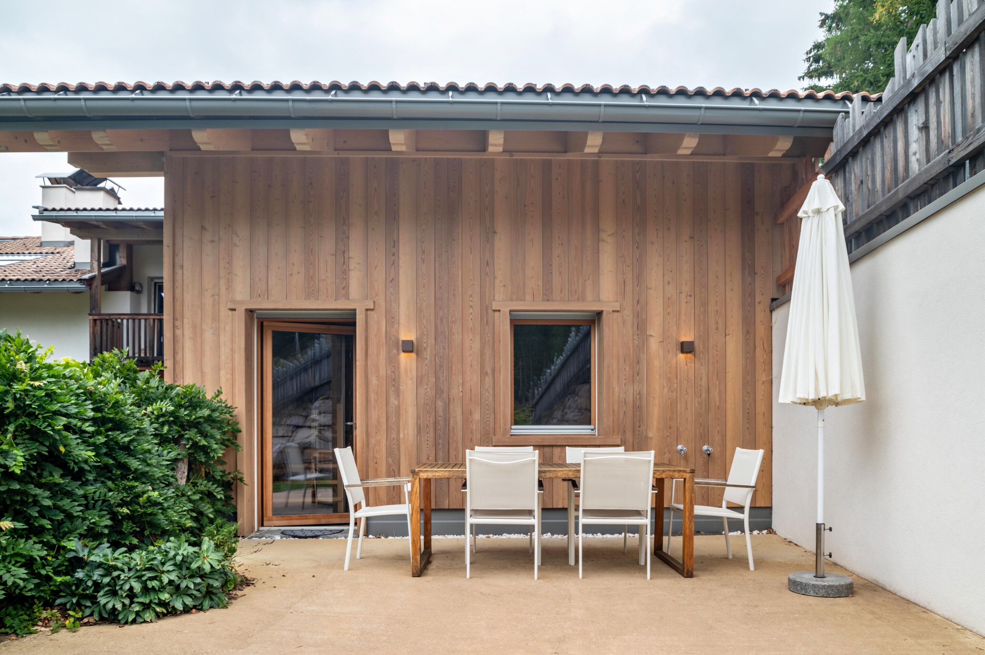 A wooden exterior wall with two windows, a covered patio area with a table and chairs, and a white umbrella.