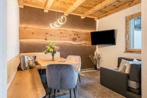 Un interior moderno con techo de madera, un televisor de pantalla plana, un banco de madera, una mesa de comedor con un florero y un sofá gris.