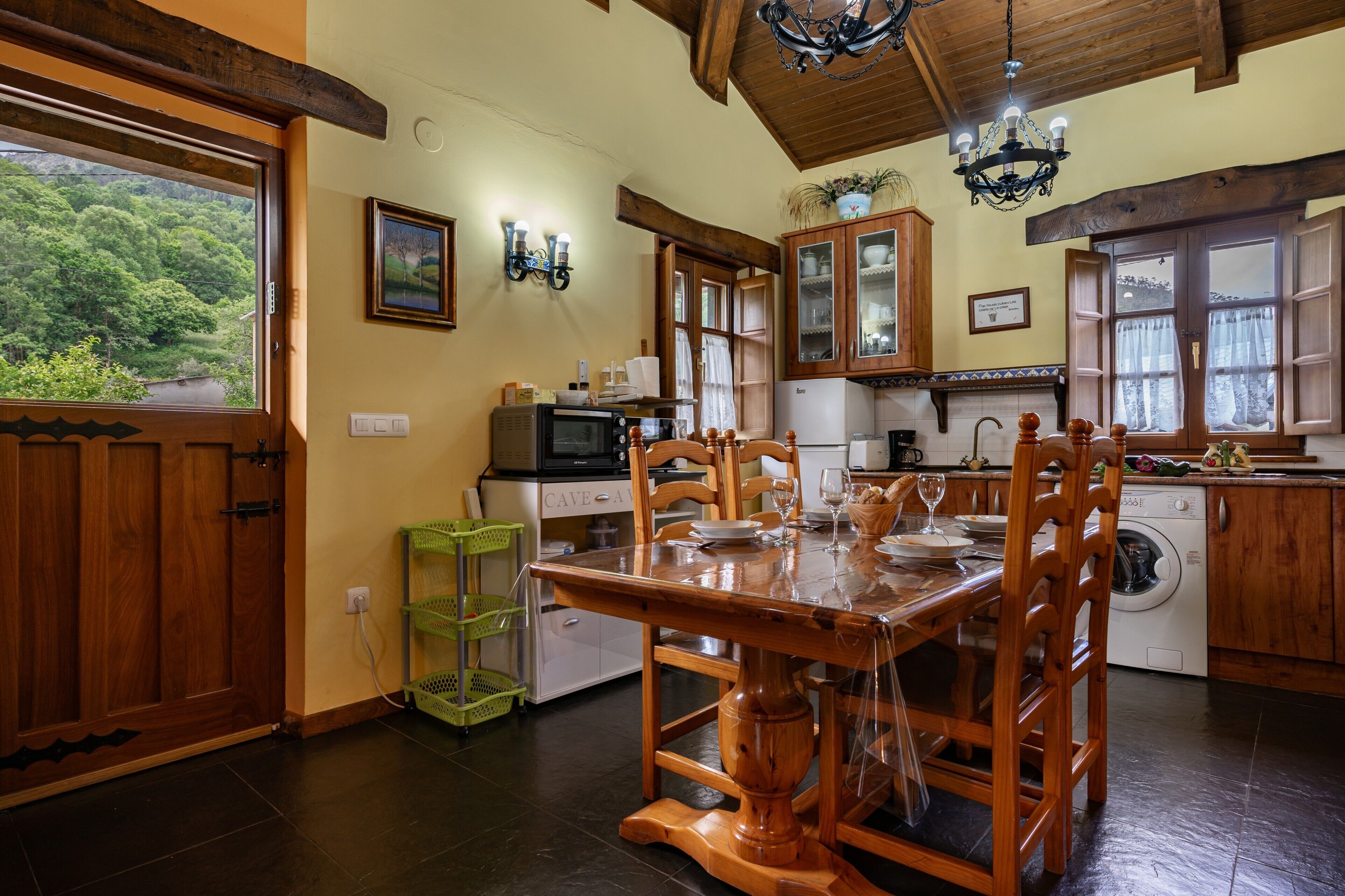 A wooden dining table with chairs, a kitchen with wooden cabinets, a washing machine, and a view of greenery outside.