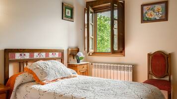 Un dormitorio con cabecera de madera, cama con colcha floral, mesita de noche, cómoda y una ventana con persiana abierta, además de un radiador.