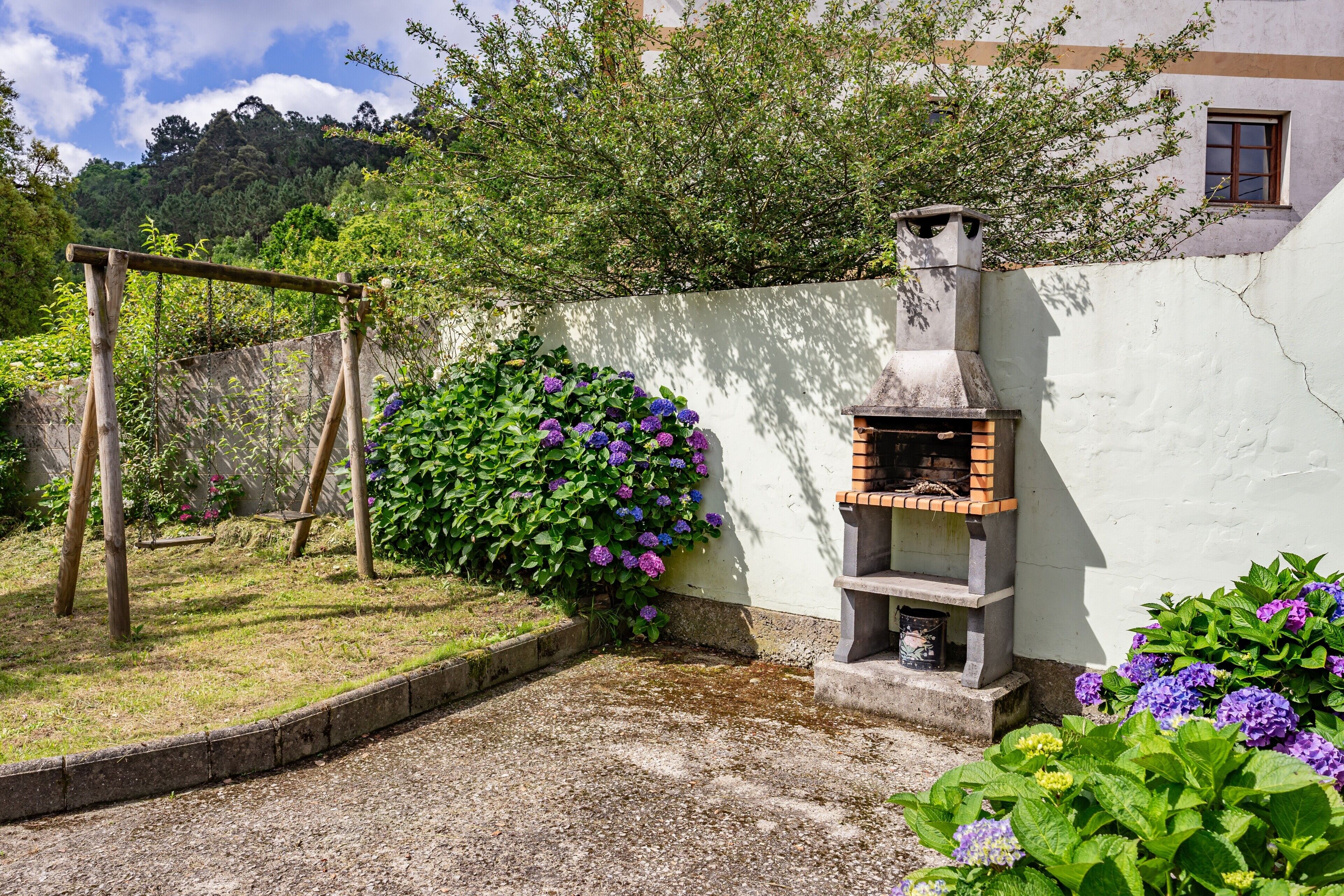 A garden with a wooden swing set, a brick oven, and flowering plants.