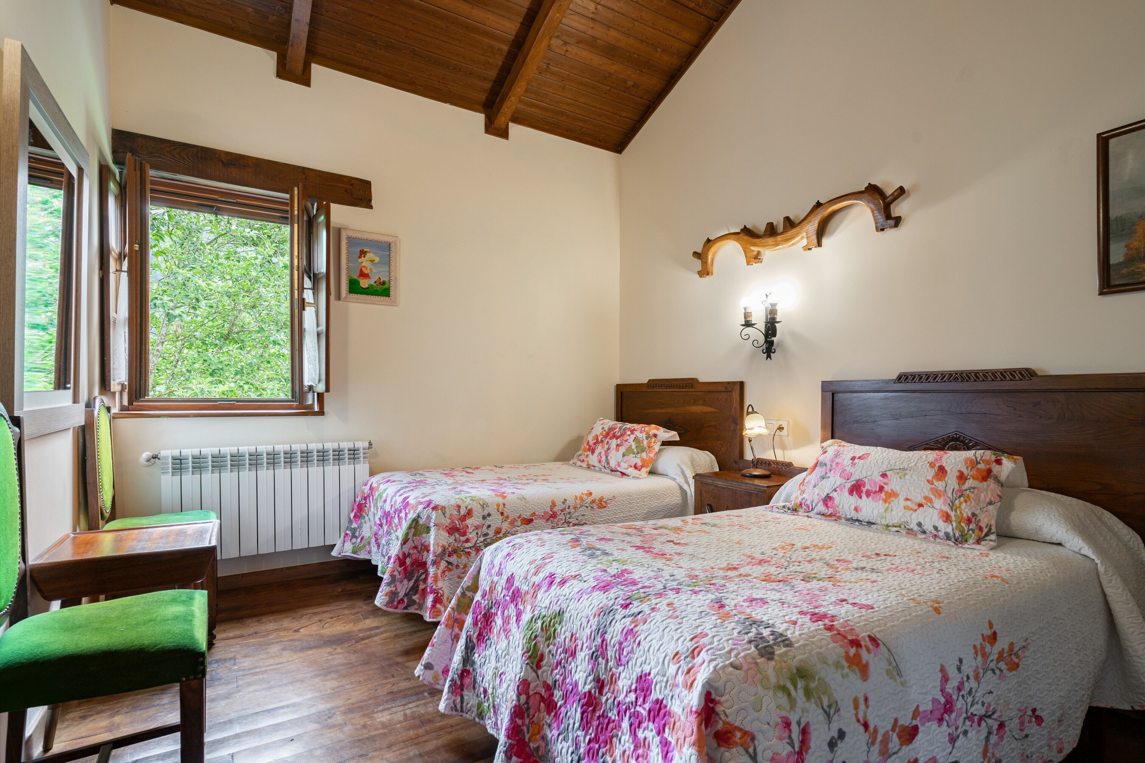 Una habitación con dos camas, una silla de madera, un radiador, una ventana con vistas a la vegetación y una lámpara fijada a la pared.