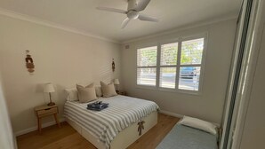 Un dormitorio con cama, mesita de noche, lámpara, ventilador de techo y ventana con persianas.
