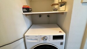 Un área de lavandería pequeña con una lavadora de carga frontal, un estante superior con algunos objetos y piso de baldosas.