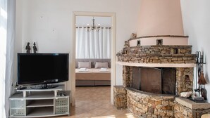 A living room with a television on a stand, a stone fireplace, and a bed with a view. - Holiday Home 'Naxos Family House In Vivlos' with Sea View, Wi-Fi and Air Conditioning (Naxos)