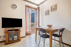 A living room with a flat-screen TV on a wooden stand, a dining table with chairs, a wooden bowl, and a view of mountains through a sliding door.