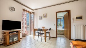 Una sala de estar con televisión, mueble bajo de madera, mesa de comedor con sillas y vista a través de una puerta abierta.
