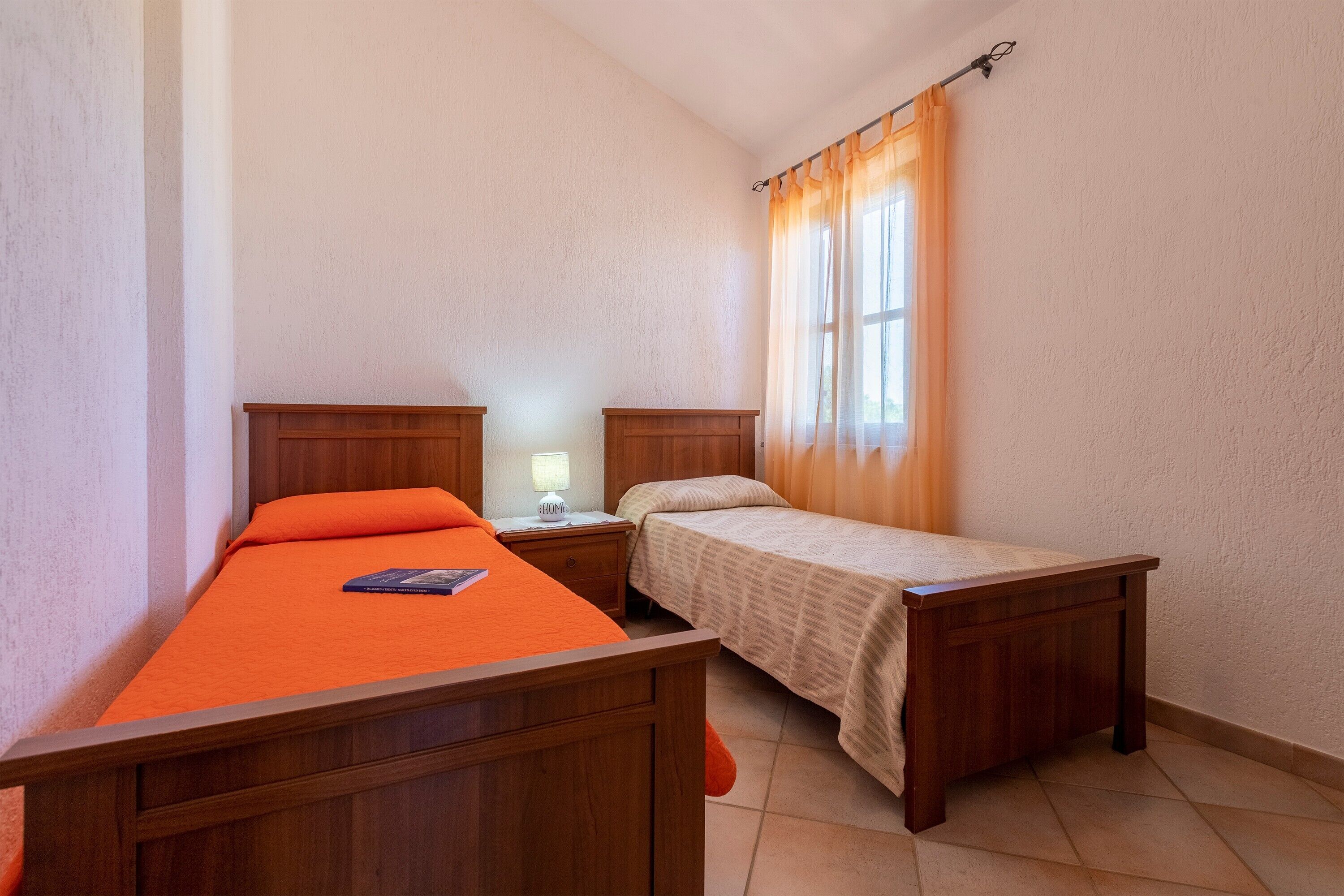 A small bedroom with two wooden beds, a nightstand, a lamp, and a window with curtains.