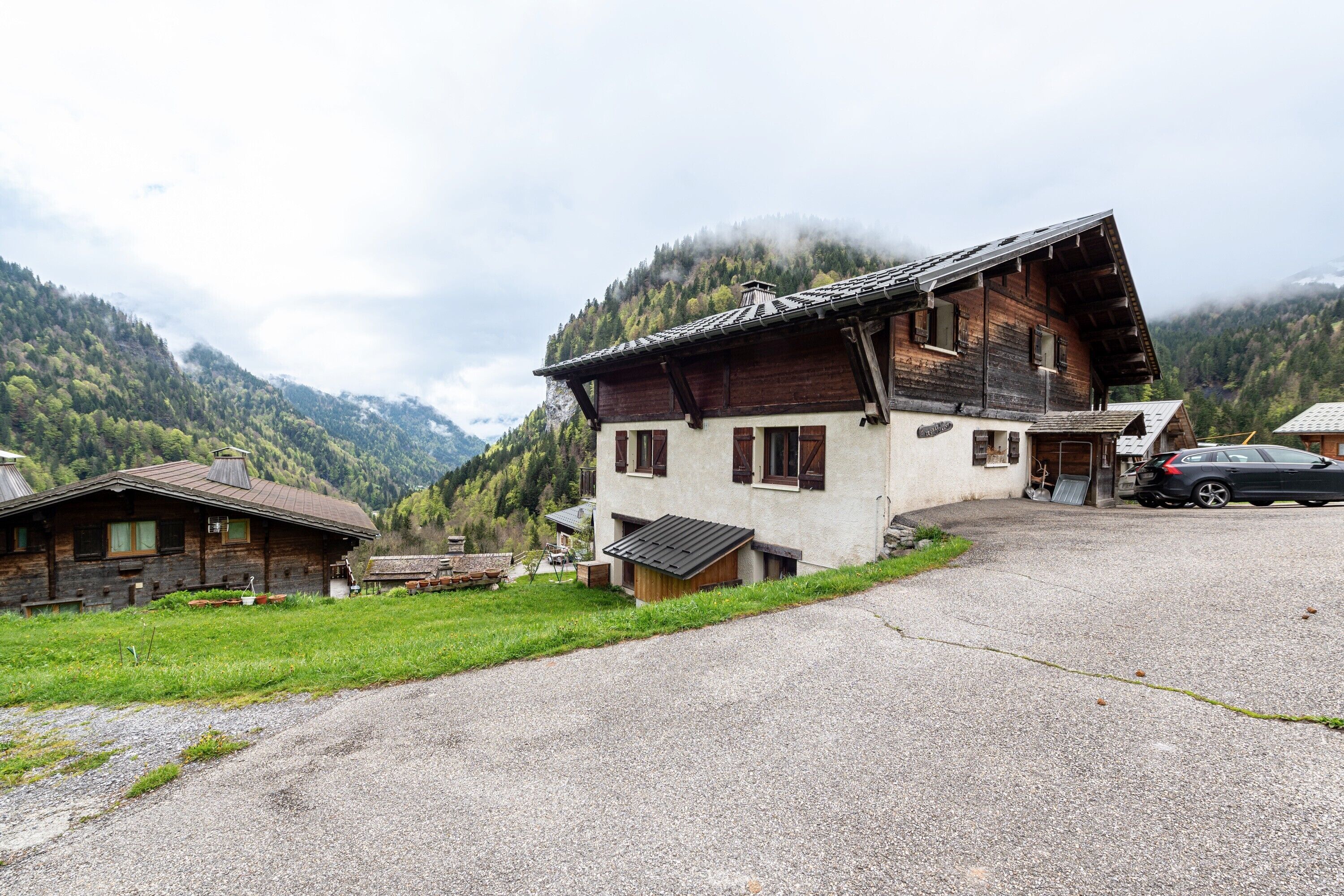 A wooden house with a sloped roof, a gravel driveway, and a mountain backdrop.