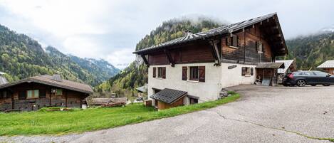 A wooden house with a sloped roof, a gravel driveway, and a mountain backdrop.