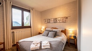 A neatly made bed with pillows and a blanket, a bedside table with a lamp, a window with curtains, and a wall with a decorative panel. - Apartment 'Chalet Le Grand Nant' with Balcony and Wi-Fi (La Giettaz)