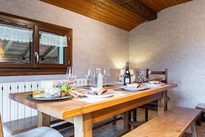 A wooden dining table set with plates, glasses, and a bottle, with a window above and a bench to the right.