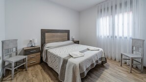 Un dormitorio con cama, dos mesitas de noche, una silla, una lámpara y una ventana con cortinas.