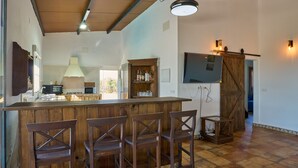 A kitchen with a wooden bar, bar stools, a cabinet with bottles, a lamp, and a TV mounted on the wall.