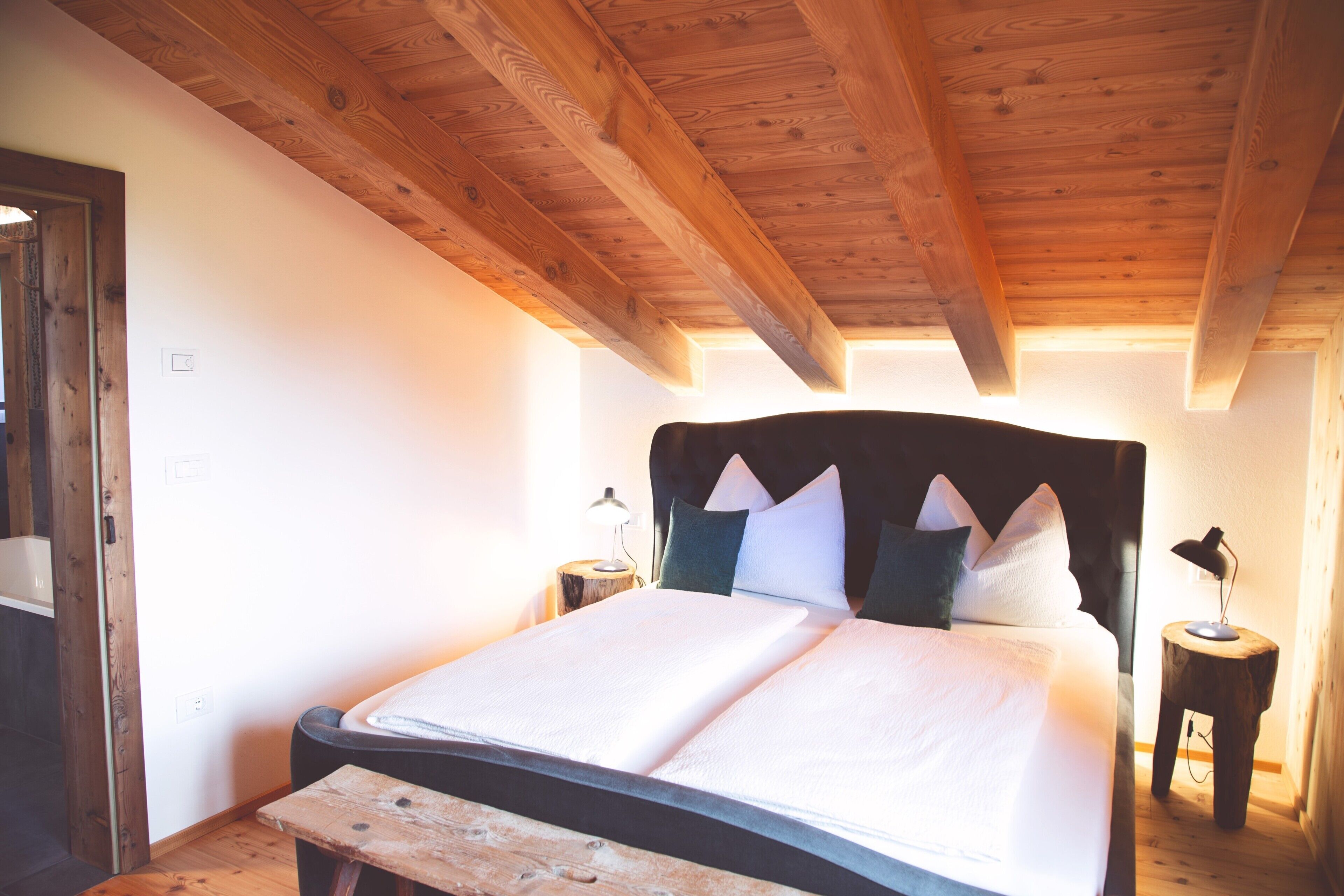 A bedroom with a wooden ceiling, a bed with white bedding, two bedside tables with lamps, and a wooden bench.