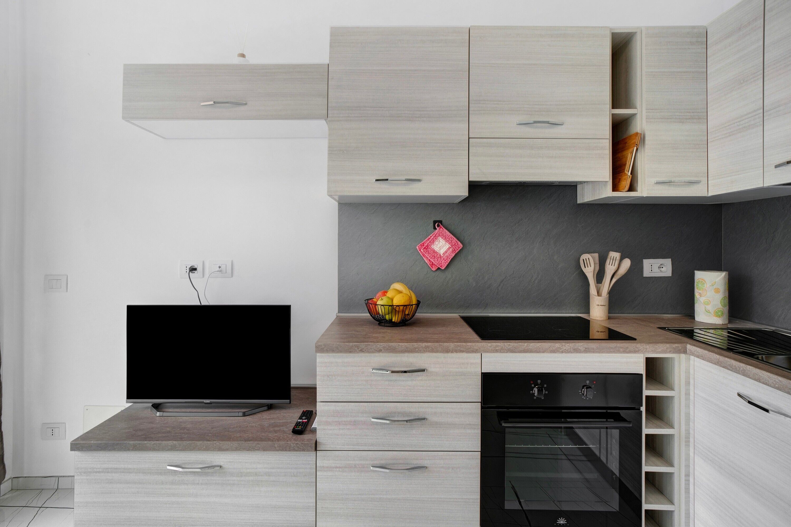Une cuisine moderne équipée d’un téléviseur à écran plat, d’un saladier rempli de fruits et d’ustensiles de cuisine en bois posés sur le plan de travail.