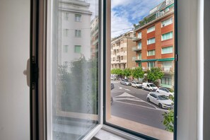 Una ventana con vistas a un edificio de varios pisos, una calle con coches estacionados y un cielo despejado.